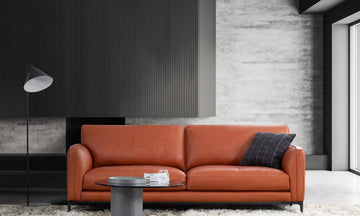 An orange leather sofa in a modern living room setting with a black floor lamp, a round glass coffee table, and a textured gray wall background.