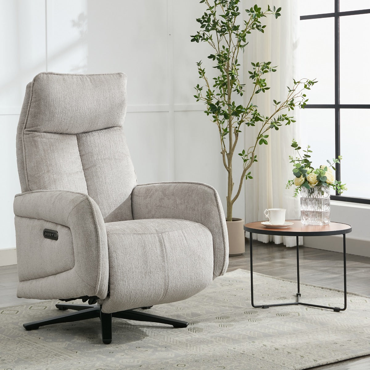 A light beige upholstered recliner chair with a black swivel base, shown in a bright living room setting next to a small round side table with a plant and vase.