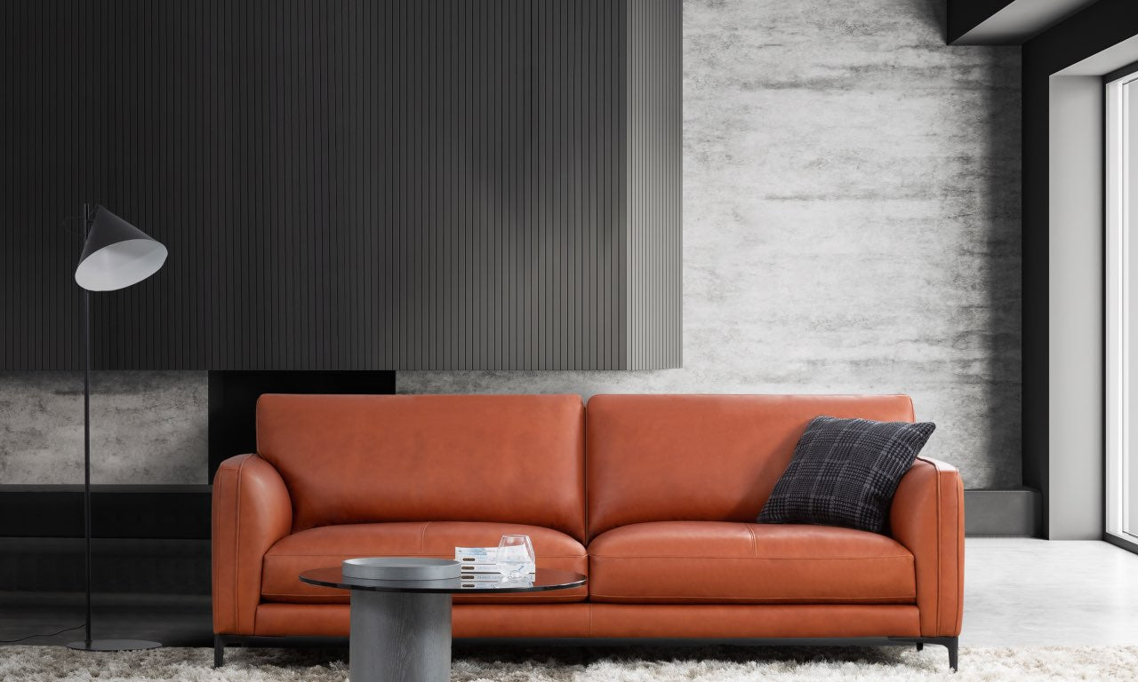 An orange leather sofa in a modern living room setting with a black floor lamp, a round glass coffee table, and a textured gray wall background.