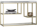 A modern console table with a brushed gold metal frame, a mirrored top surface, and two glass shelves. The left shelf holds a stack of books and a dark bowl, while the right side features a hanging glass candle holder.