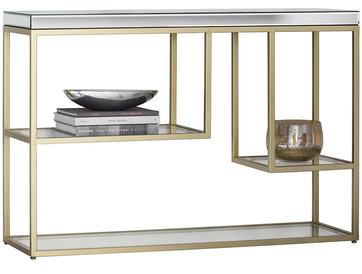 A modern console table with a brushed gold metal frame, a mirrored top surface, and two glass shelves. The left shelf holds a stack of books and a dark bowl, while the right side features a hanging glass candle holder.