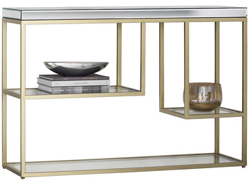 A modern console table with a brushed gold metal frame, a mirrored top surface, and two glass shelves. The left shelf holds a stack of books and a dark bowl, while the right side features a hanging glass candle holder.