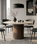 A modern round dining table with a light wood top and a dark wood pedestal base, surrounded by four gray upholstered chairs in a contemporary dining room setting.