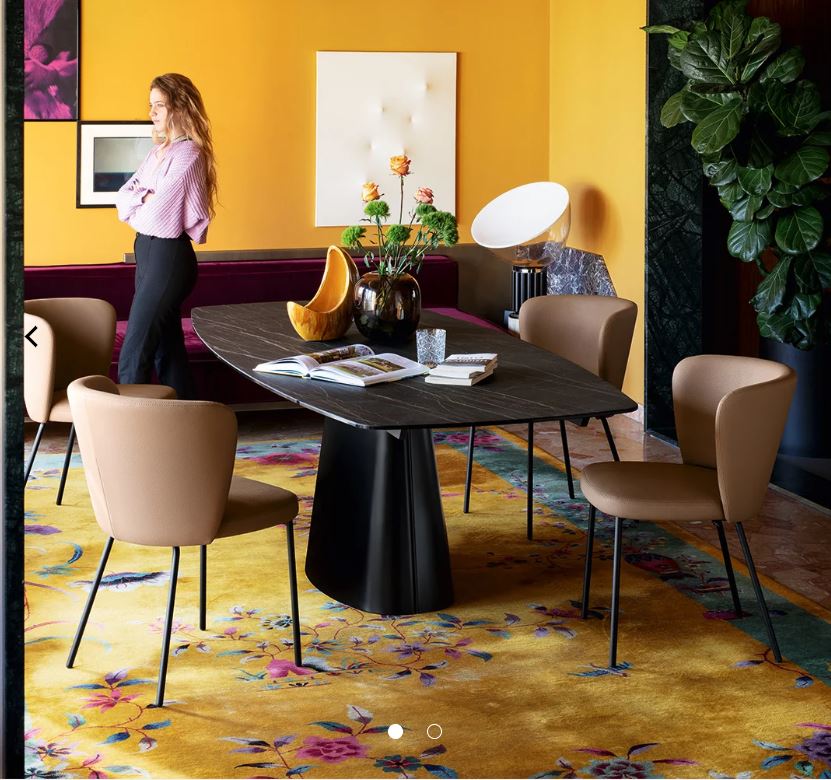 A dining room scene with a dark rectangular table and four beige upholstered chairs, surrounded by a yellow floral rug, mustard-yellow walls, and a woman standing in the background near a sofa and decor.